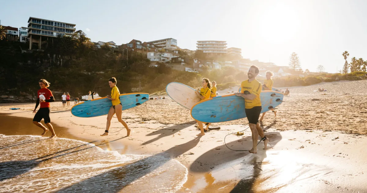 Group of surf students in yellow rash vests carrying surfboards along Freshwater Beach at sunrise, attending a lesson with Manly Surf School.