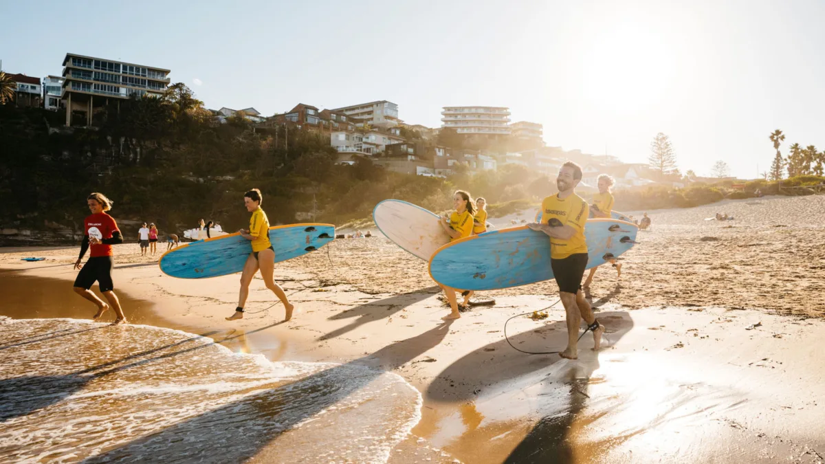 Group of surf students in yellow rash vests carrying surfboards along Freshwater Beach at sunrise, attending a lesson with Manly Surf School.