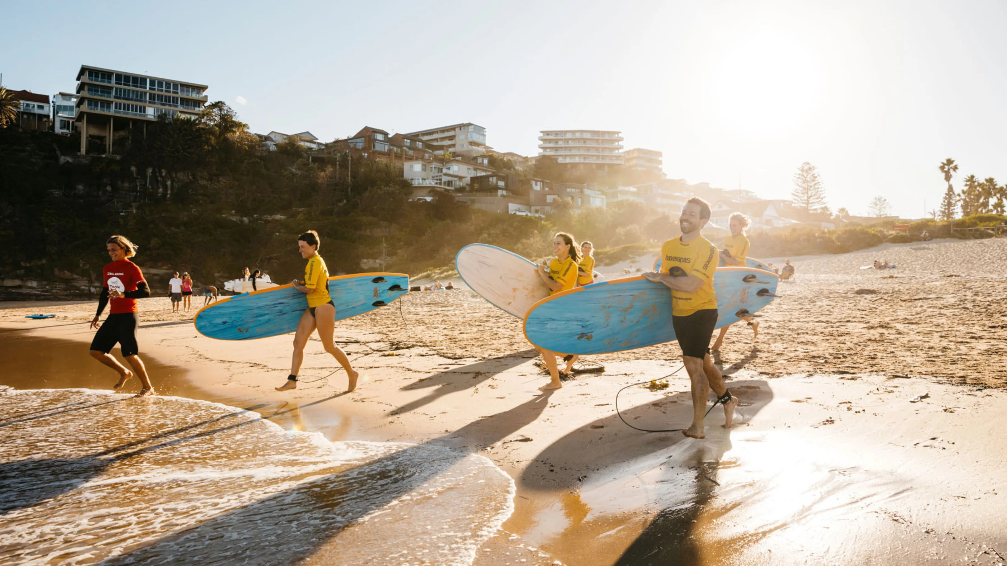 Group of surf students in yellow rash vests carrying surfboards along Freshwater Beach at sunrise, attending a lesson with Manly Surf School.