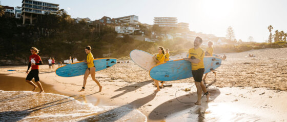 Group of surf students in yellow rash vests carrying surfboards along Freshwater Beach at sunrise, attending a lesson with Manly Surf School.