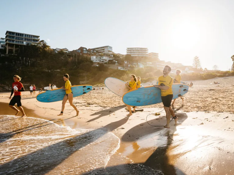 Group of surf students in yellow rash vests carrying surfboards along Freshwater Beach at sunrise, attending a lesson with Manly Surf School.