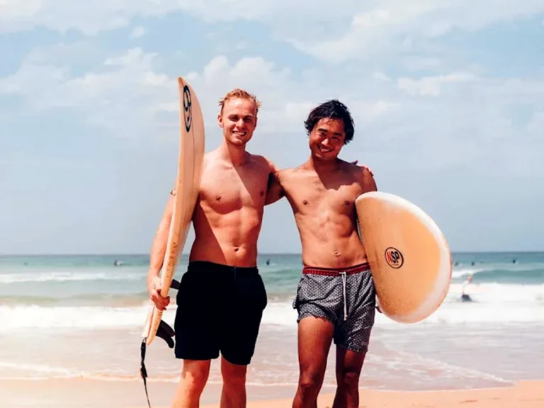 Two young male surfers smiling with surfboards under their arms, standing on Manly Beach with waves and blue sky in the background.
