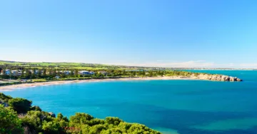 A scenic view of Victor Harbor's coastline with clear blue waters, sandy beaches, coastal homes, and green rolling hills under a bright blue sky.
